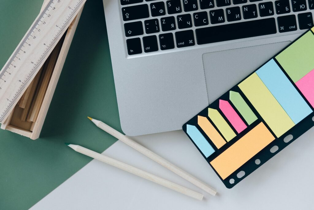 Flat lay of a workspace with a silver laptop, colorful sticky notes, two white pencils, and a wooden ruler box on a green and white surface