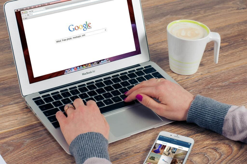 Hands typing on a MacBook Air with Google search open, a cup of coffee beside the laptop and a smartphone on the table